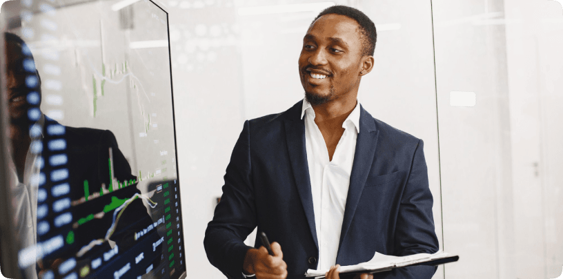 A man in a suit smiling beside a screen of currency charts